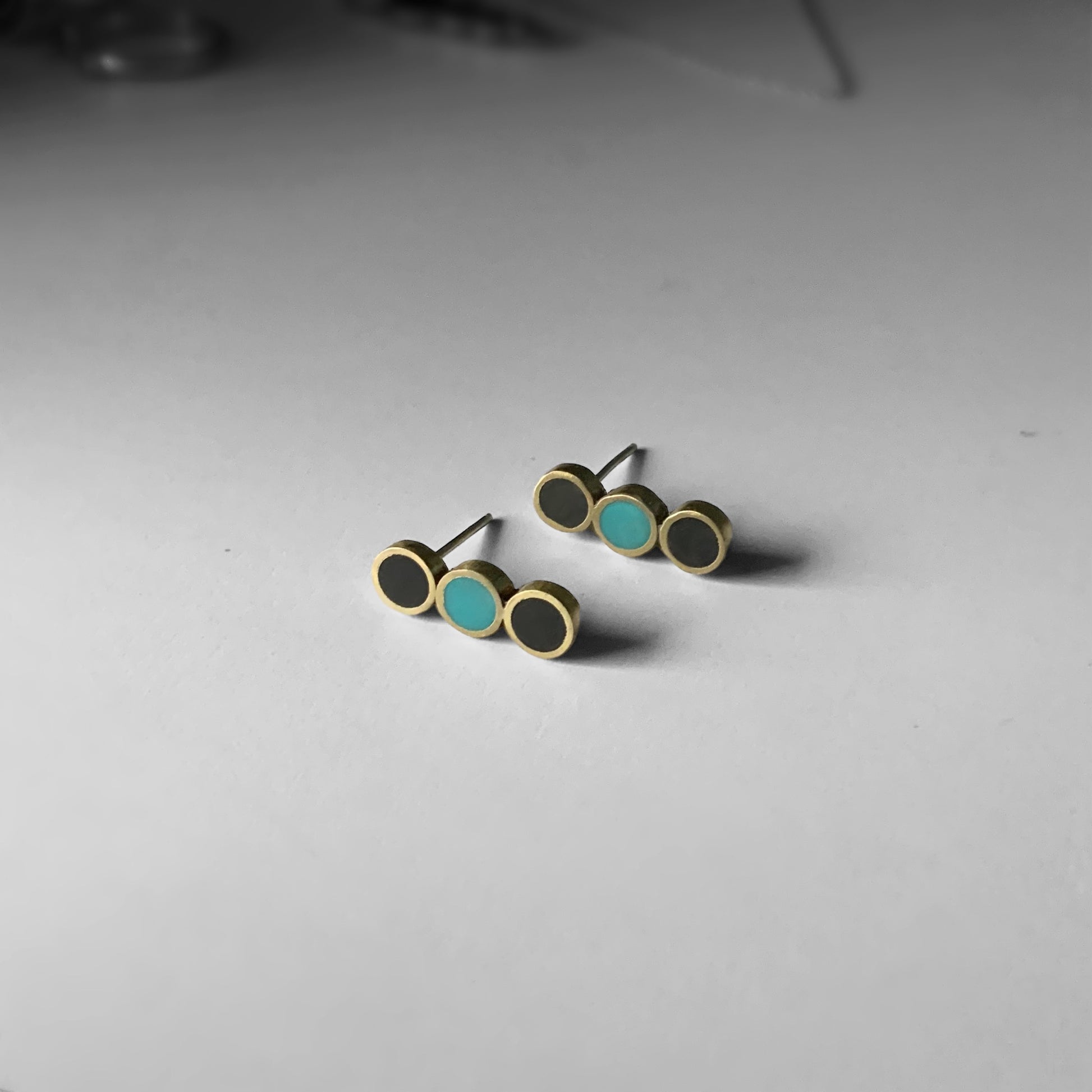 A pair of brass stud earrings with three circular accents—black and turquoise—arranged in a minimalist linear design, displayed on a light gray surface with subtle background details
