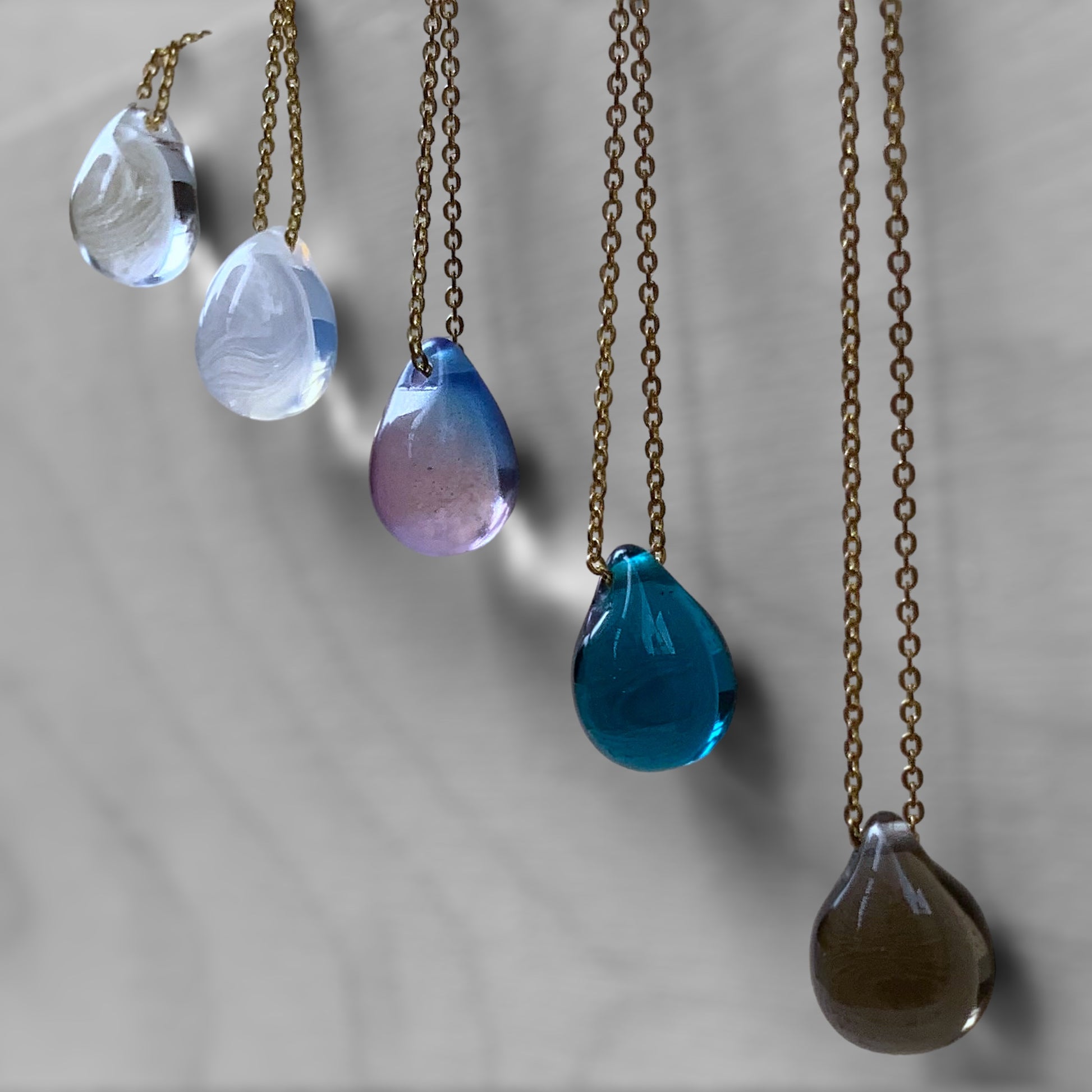 A collection of handcrafted necklaces featuring gradient translucent teardrop glass pendants in shades of white, purple, aqua blue, and brown, suspended on brass chains and displayed on a light gray background.
