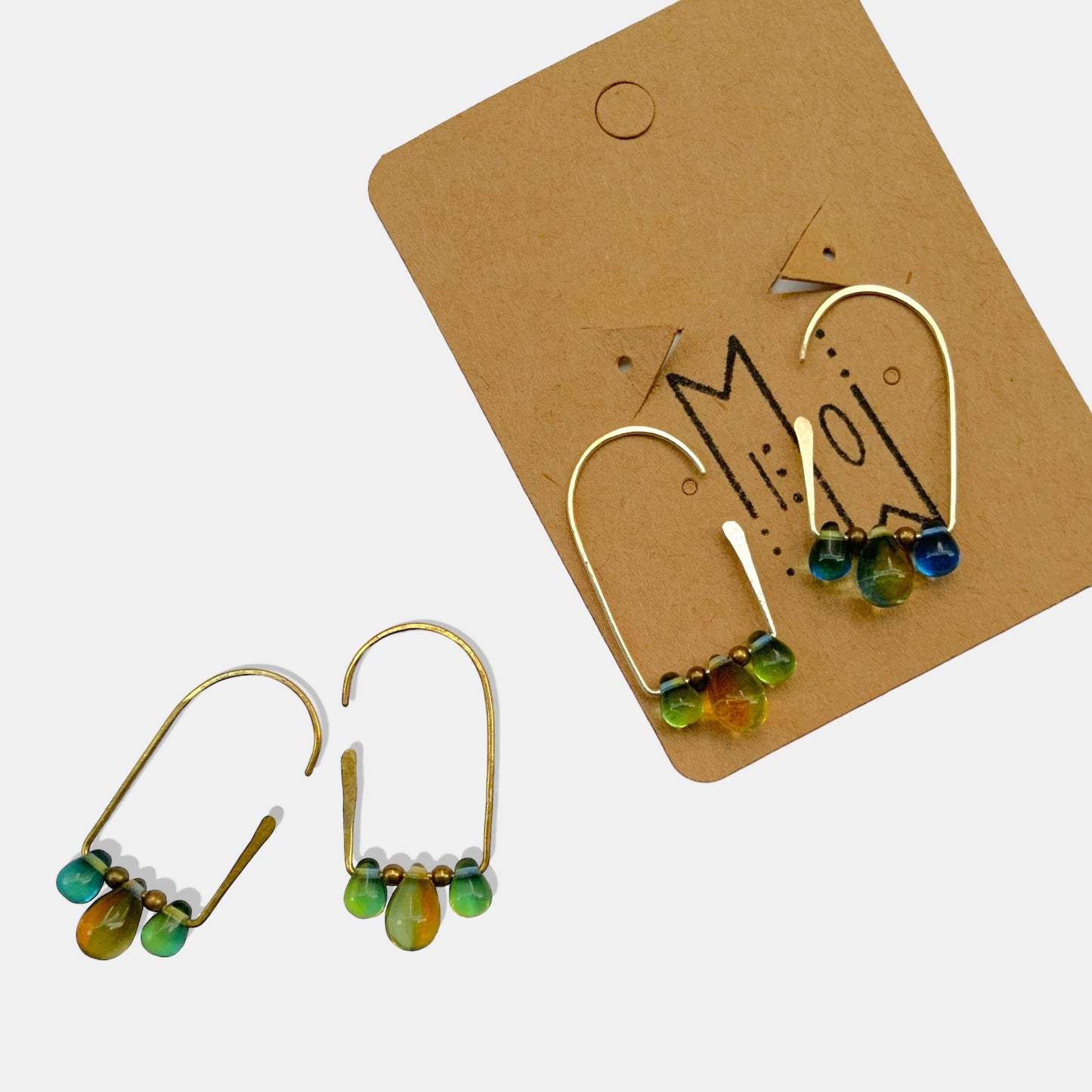 A pair of gradient silver and gold teardrop hoop earrings in shades of green, amber, and blue, displayed alongside branded Jewelry by Meow packaging for an elegant presentation.