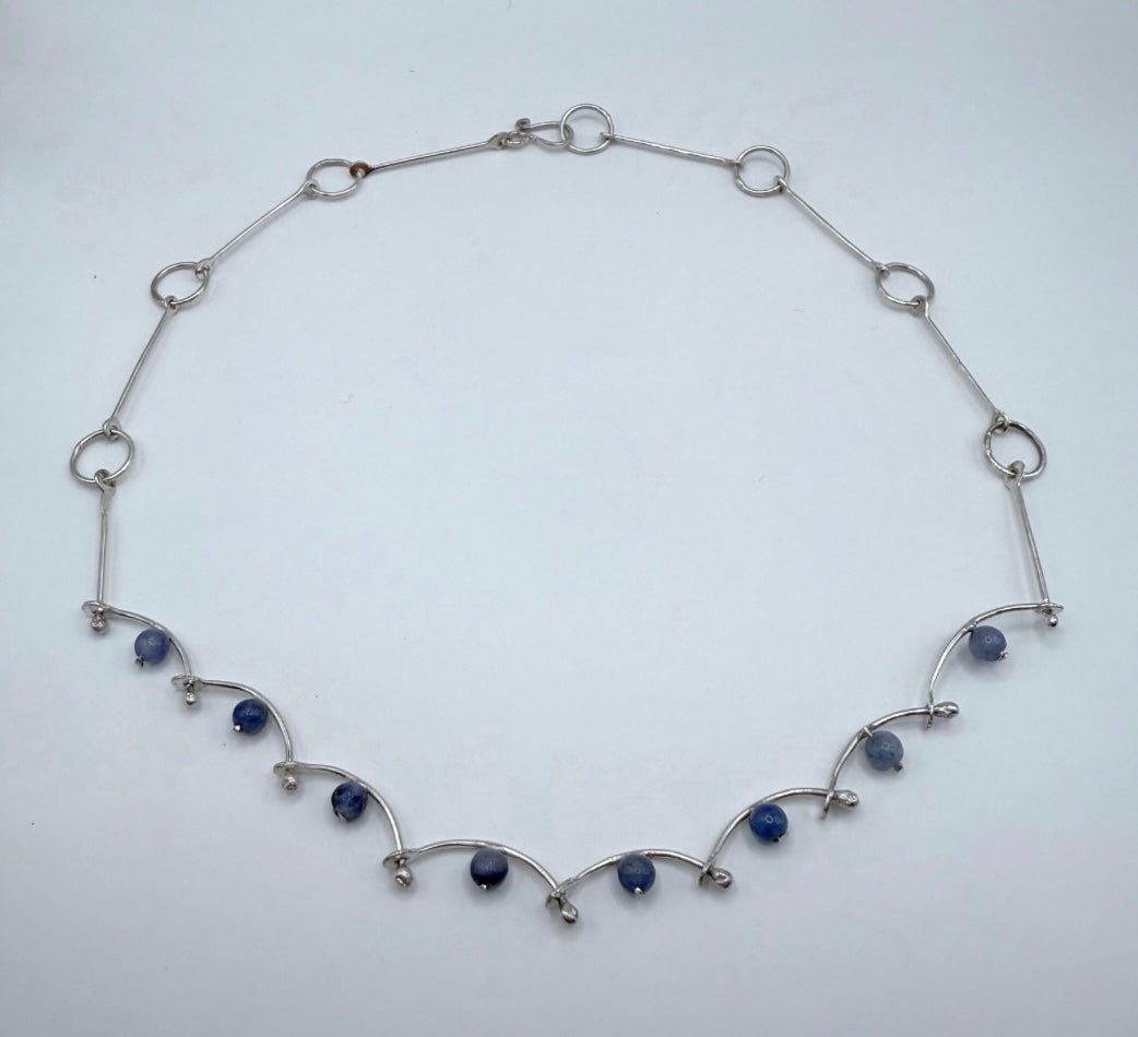 Overhead view of a handmade sterling silver necklace with blue aventurine beads and curved, wave-like hammered links, minimalist and organic Greek jewelry.
