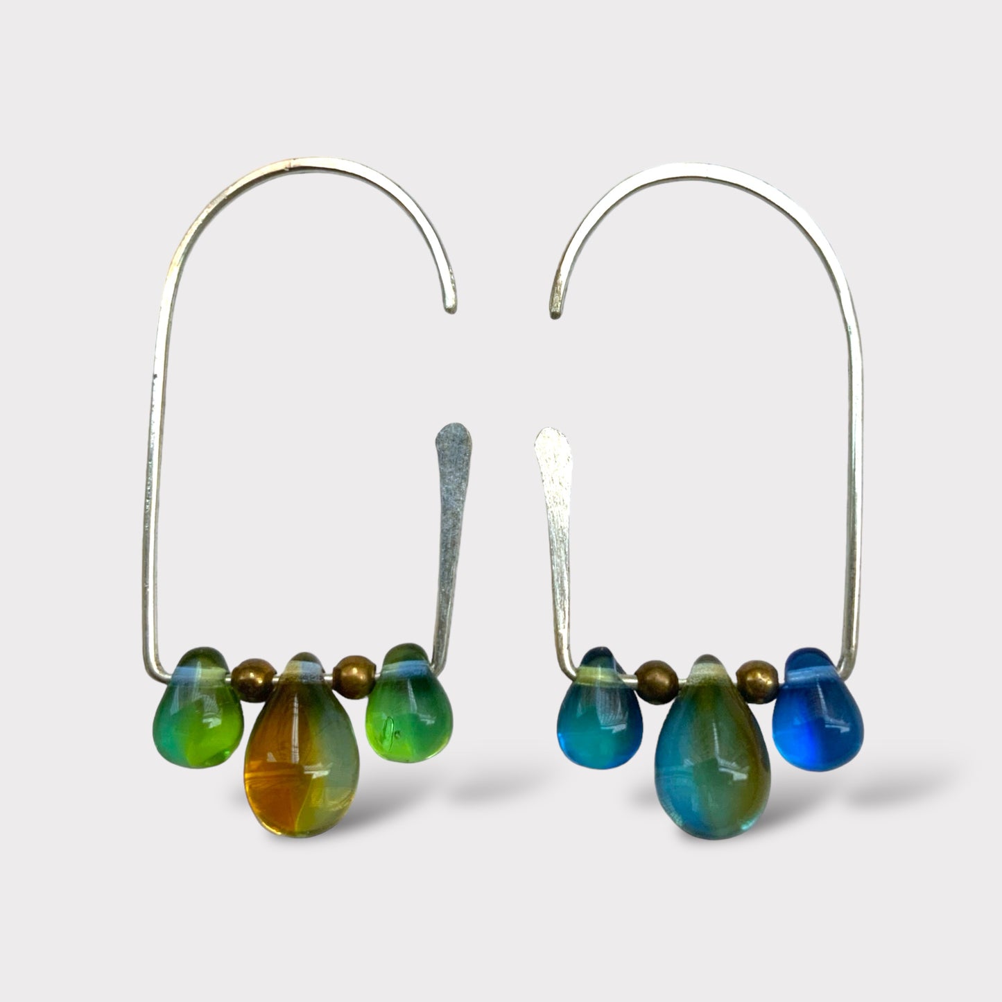 A pair of handcrafted silver hoop earrings featuring gradient teardrop glass pendants in shades of green, blue, and amber, accented with brass beads, displayed on a clean white background.