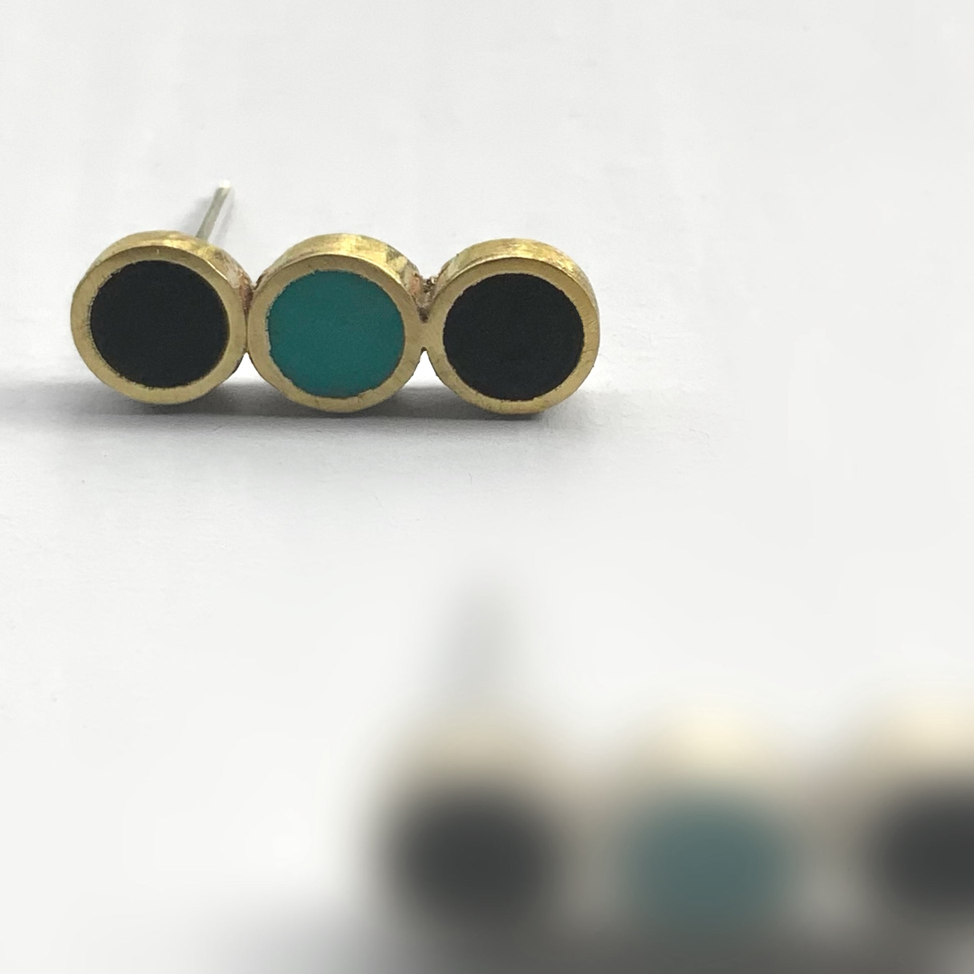 A close-up of brass stud earrings with three circular inlays, two black and one turquoise, arranged in a sleek row, styled on a white background to emphasize their geometric design.
