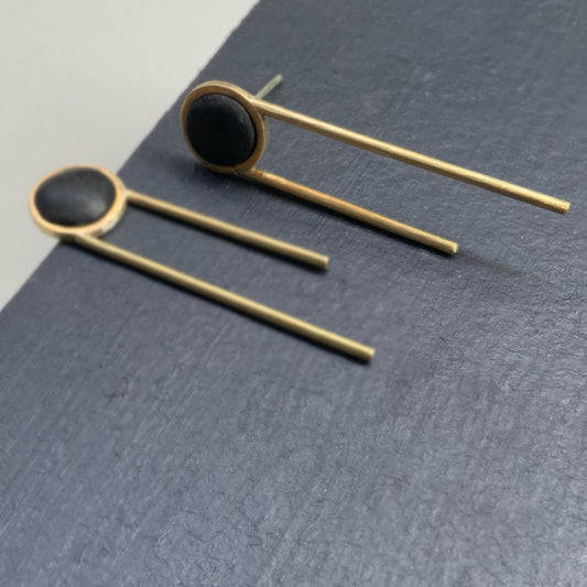 A close-up view of minimalist drop earrings featuring a circular black centerpiece set in brass with two parallel brass rods extending downward, displayed on a dark slate surface