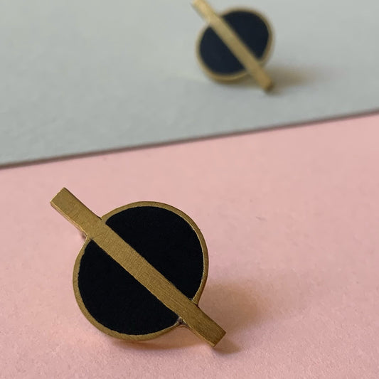 Elegant brass earrings with bold black circular details and sleek brass rods in a vertical alignment, photographed on a soft pink and gray background for contrast