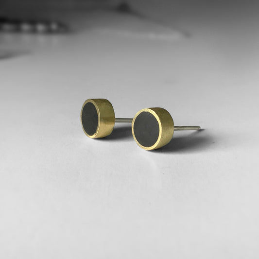 A close-up of brass stud earrings with black circular inlays, displayed on a light gray surface to emphasize their sleek and modern design.