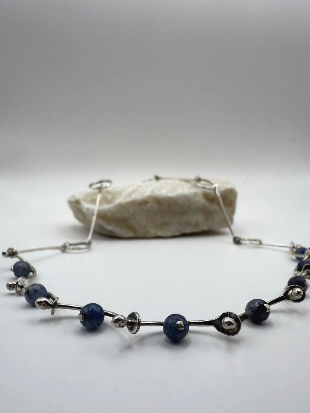 Handcrafted sterling silver necklace with blue aventurine beads and hammered details, arranged in organic, wave-inspired lines, displayed in front of a natural stone.
