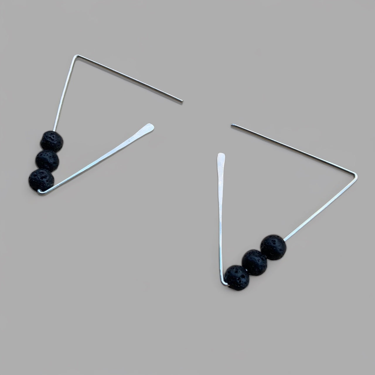 A pair of geometric silver earrings adorned with three black lava beads, styled on a light gray background. Perfect for adding a minimalist yet edgy touch to your look.