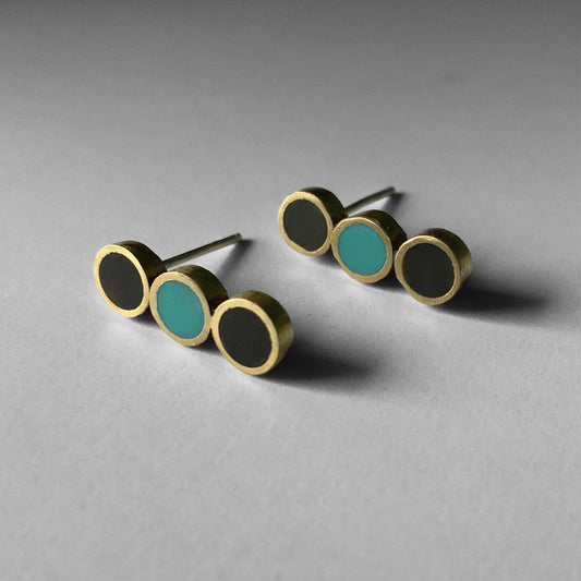 A pair of handcrafted brass stud earrings featuring three circular accents, two black and one turquoise in a linear arrangement, displayed on a light gray surface for a modern look