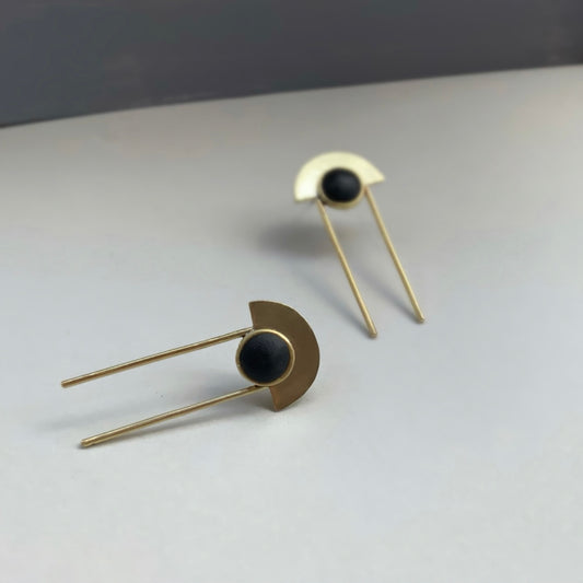 Elegant drop earrings featuring a black circular centerpiece framed by a brass half-moon, paired with long minimalist brass rods, displayed on a gradient gray surface.