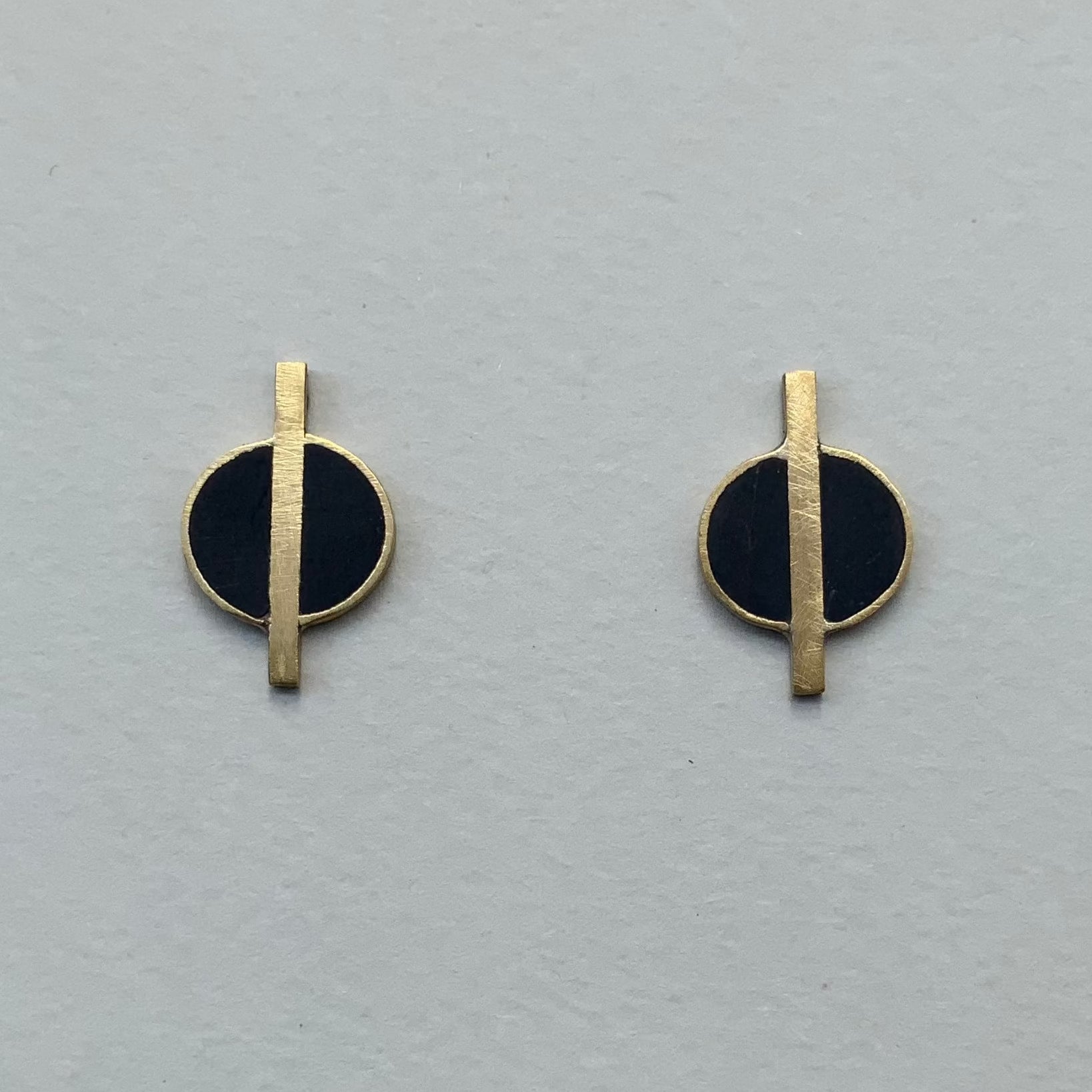 A pair of minimalist brass earrings featuring black circular accents with a vertical brass rod running through the center, displayed on a light gray background