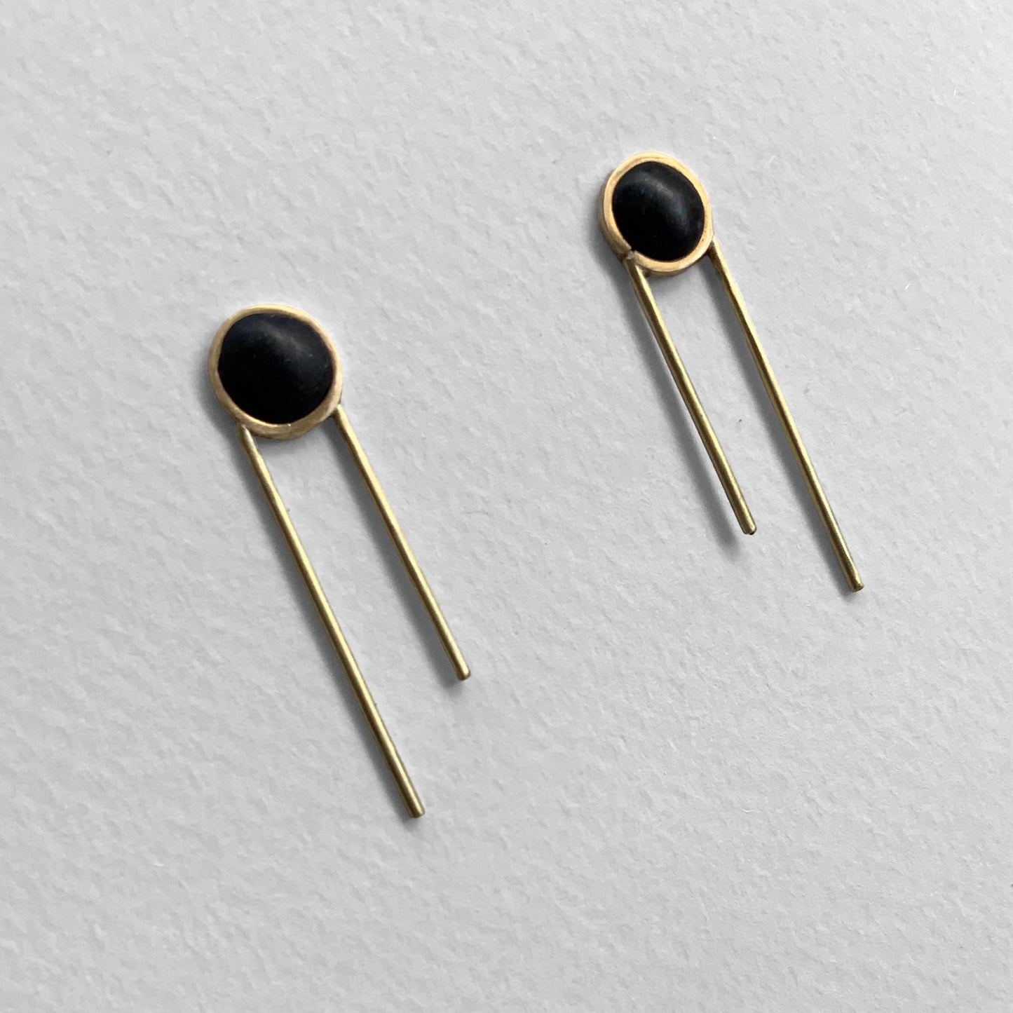 Front view of a pair of modern minimalist earrings with a black circular design encased in brass, paired with two straight brass rods, artistically arranged against a textured white background