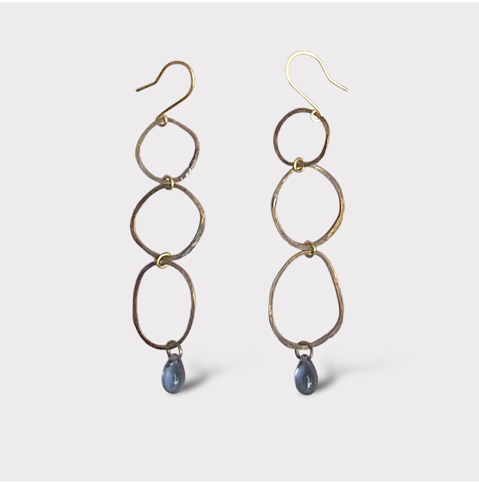 A pair of handcrafted earrings featuring interconnected oval brass links with a blue teardrop glass pendant at the bottom, suspended on gold hooks, styled on a white background for a minimalist look.