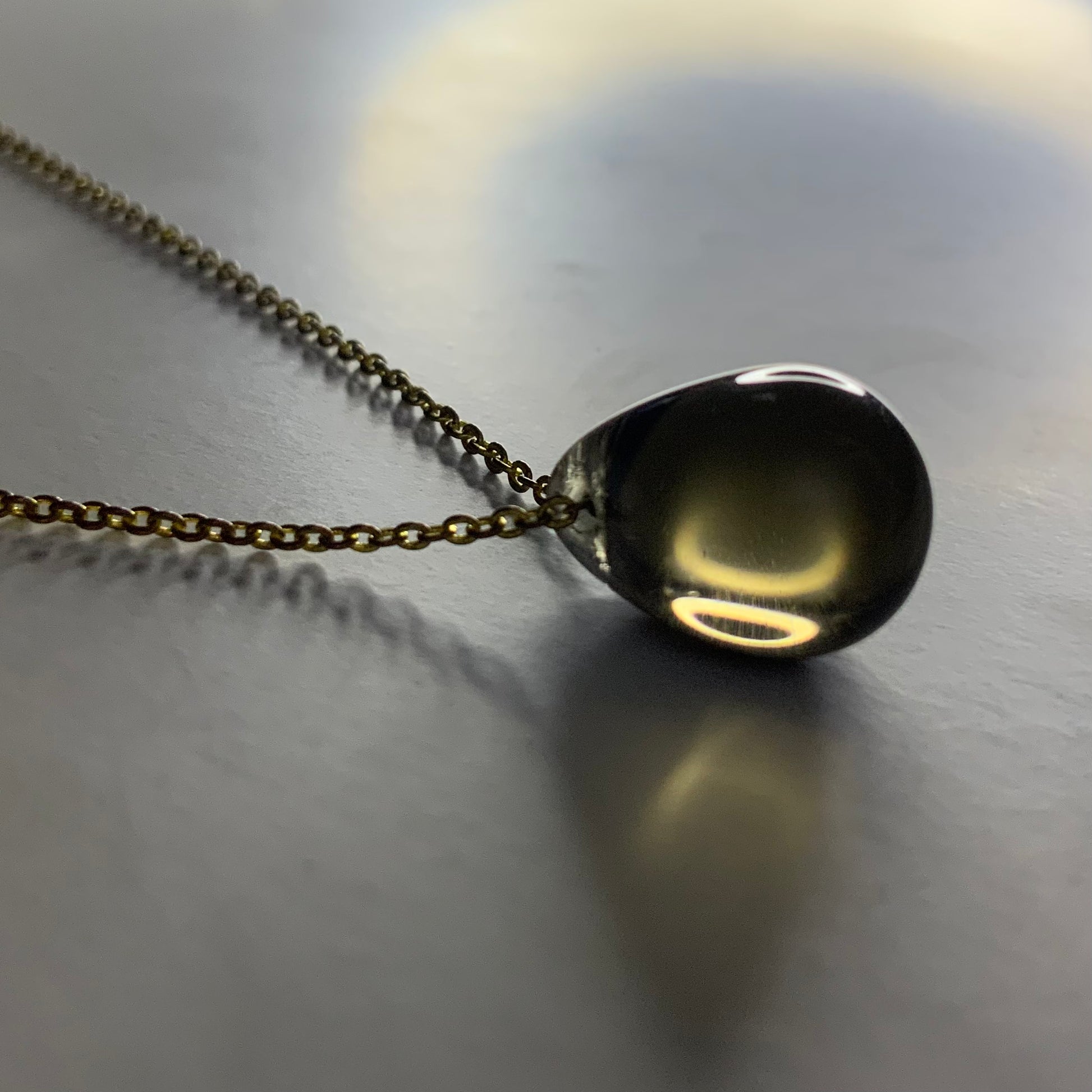 A handcrafted necklace featuring a smoky gray translucent teardrop glass pendant, elegantly suspended on a brass chain, styled on a softly lit surface for a modern and sophisticated look.