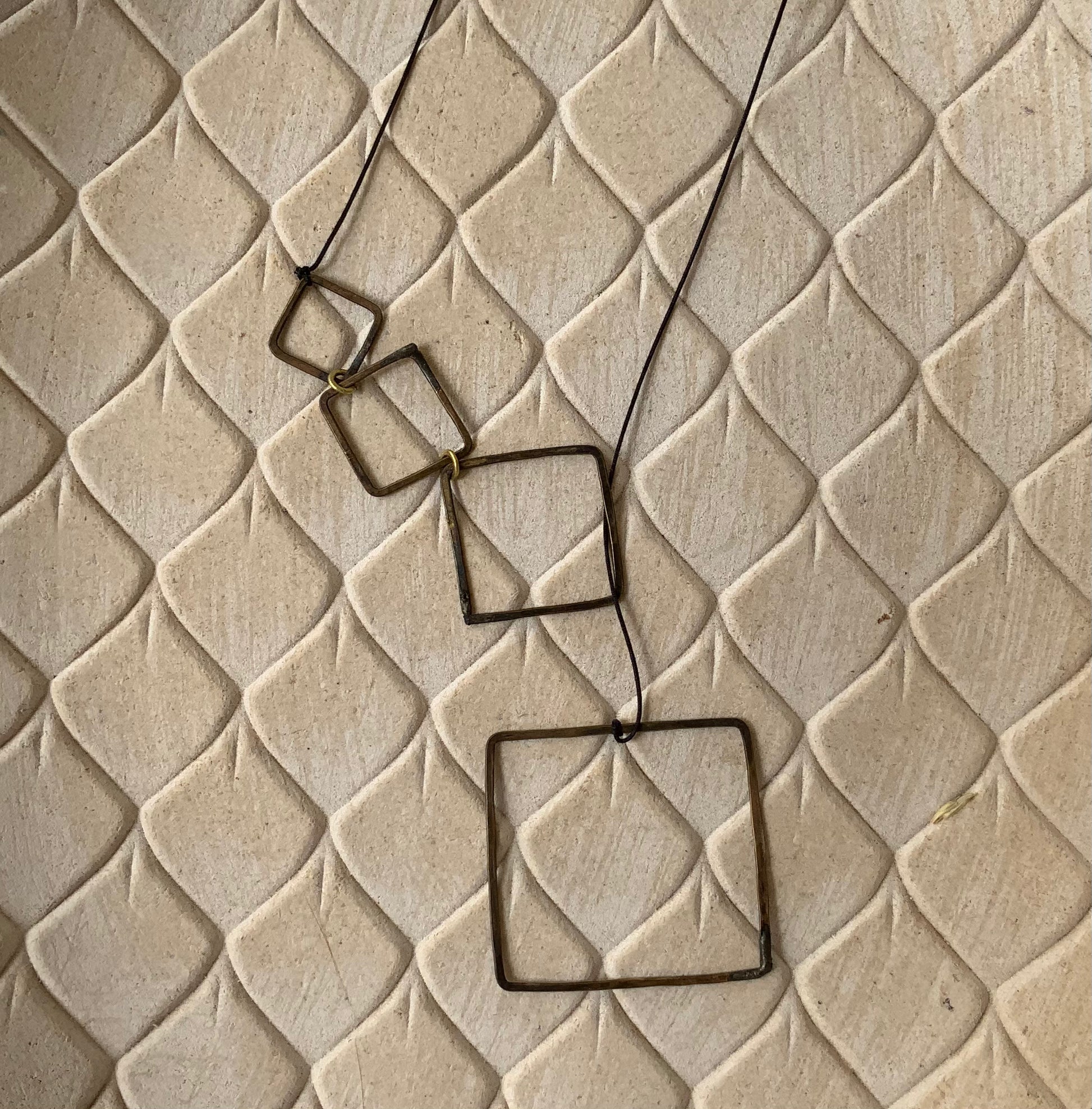 A brass lariat with cascading open square accents, styled on a beige textured background to emphasize its modern and artistic aesthetic.