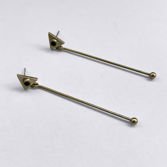Minimalist brass earrings featuring a geometric triangle stud with a black circular accent, paired with a long, sleek brass rod ending in a small ball detail, displayed on a light background