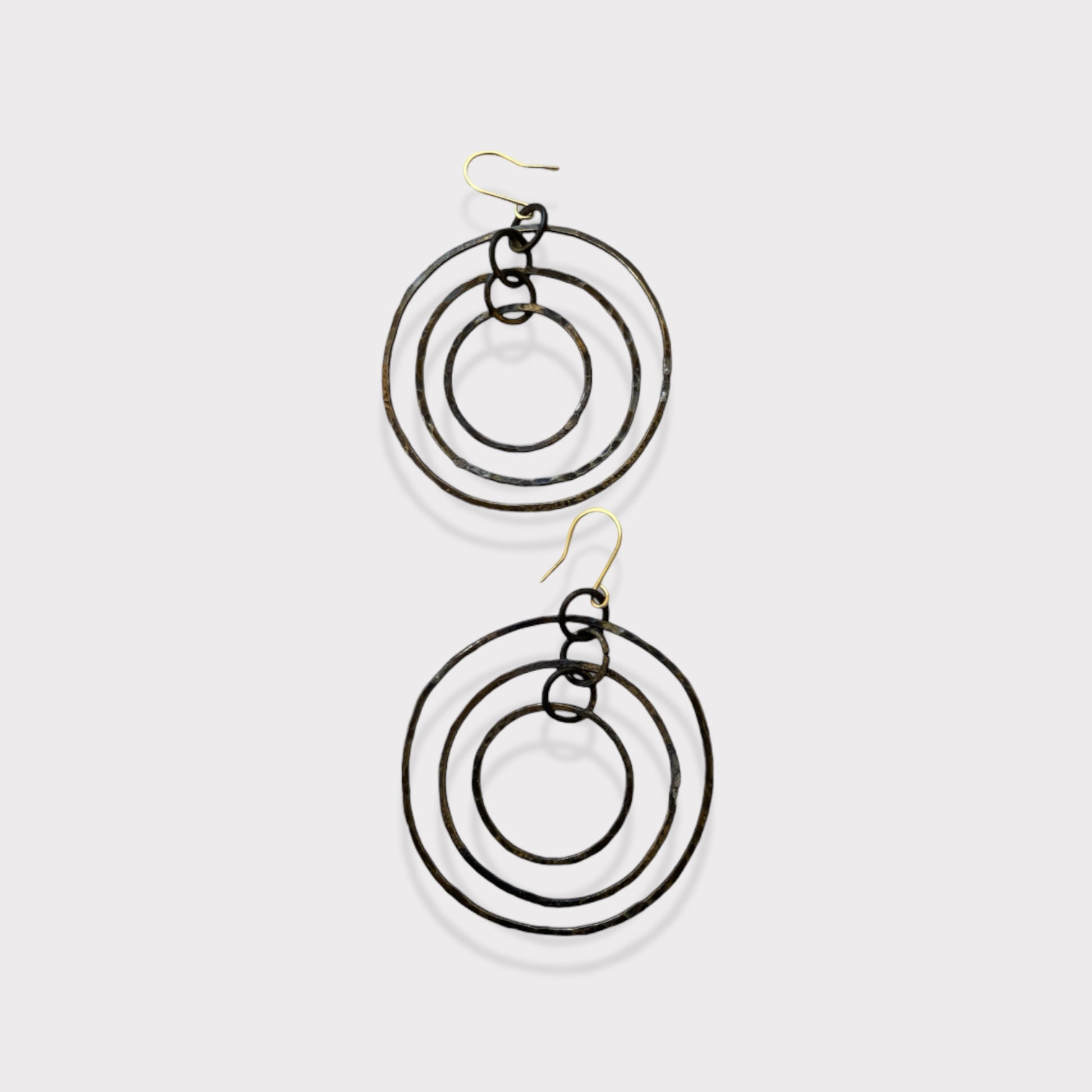 A pair of handcrafted earrings featuring three concentric hammered brass circles, suspended on gold hooks, styled on a white background for a minimalist and bold design.