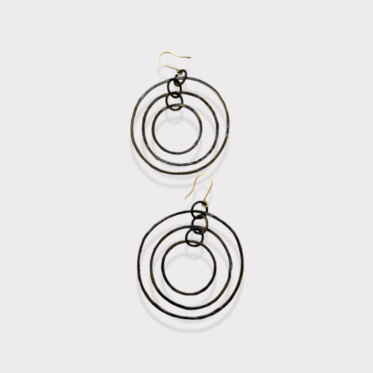 A pair of handcrafted earrings featuring three concentric hammered brass circles, suspended on gold hooks, styled on a white background for a minimalist and bold design.