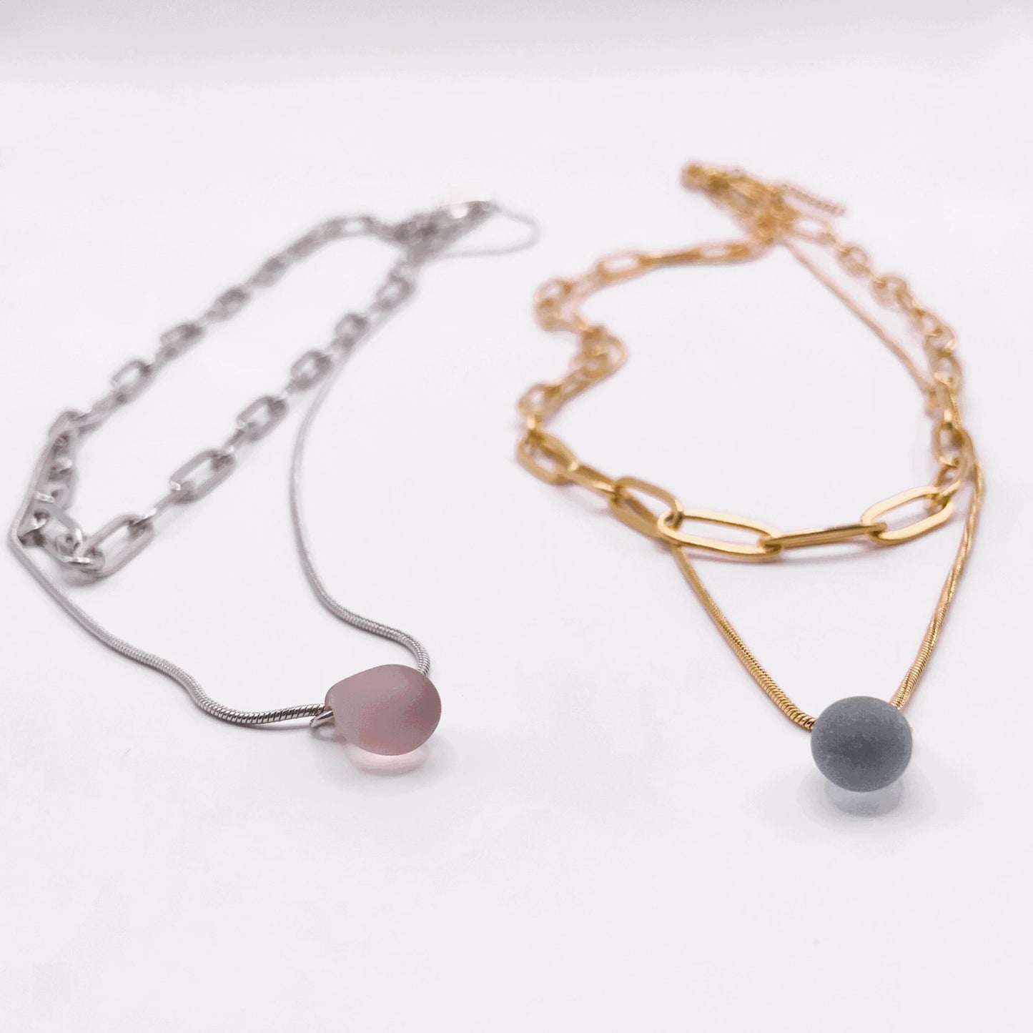 Side-by-side silver and gold chain necklaces, each featuring a frosted glass teardrop pendant—lavender on silver and blue on gold. Handcrafted in Greece, these minimalist designs combine elegance and versatility for everyday wear.