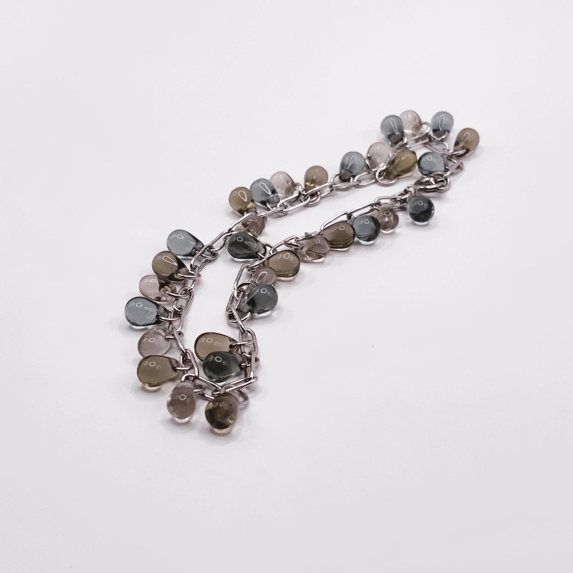 Silver chain bracelet with glass teardrop charms in earthy, blue and translucent hues, shown in a coiled arrangement on a white background.