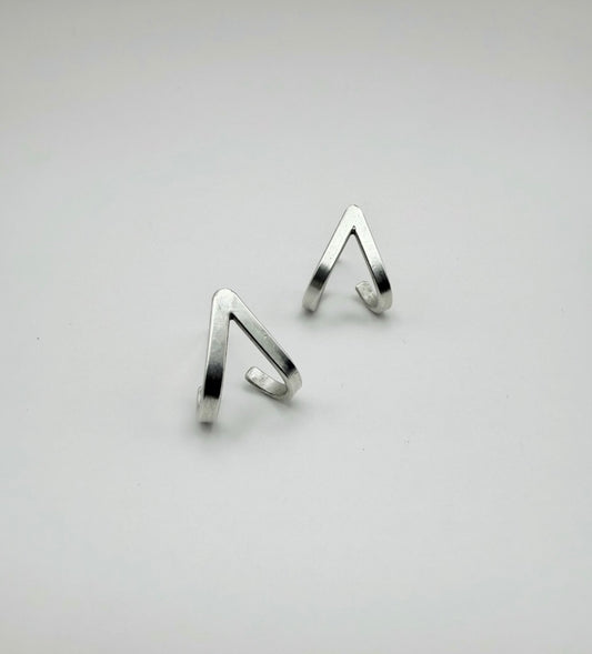 Pair of minimalist sterling silver lobe-hugger stud earrings in a chevron V shape, designed to hug the bottom of the earlobe, on a light gray background.