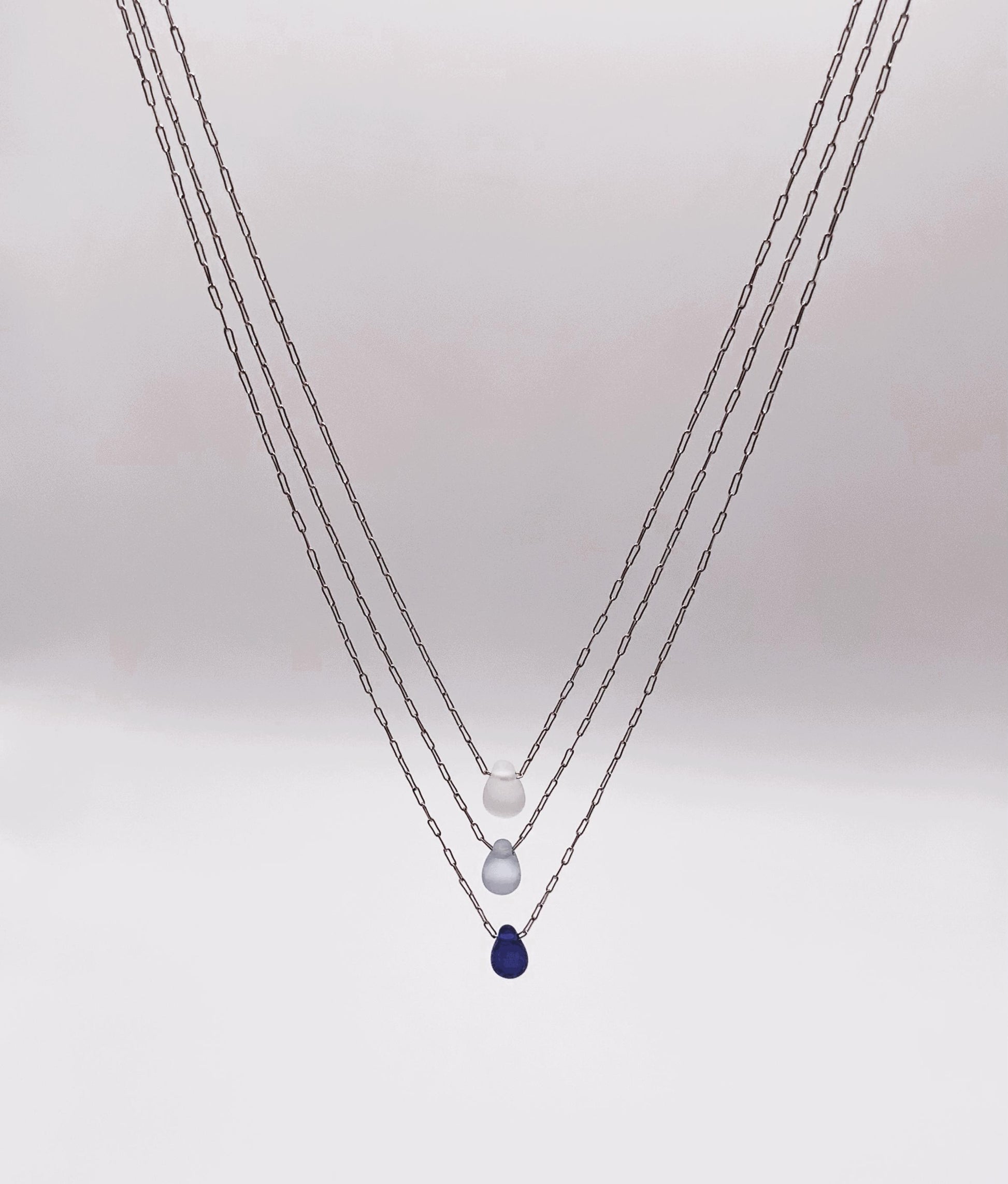 Triple-layered silver chain necklaces with frosted glass teardrop pendants in white, light blue, and deep blue. A minimalist design handcrafted for a modern, elegant look.