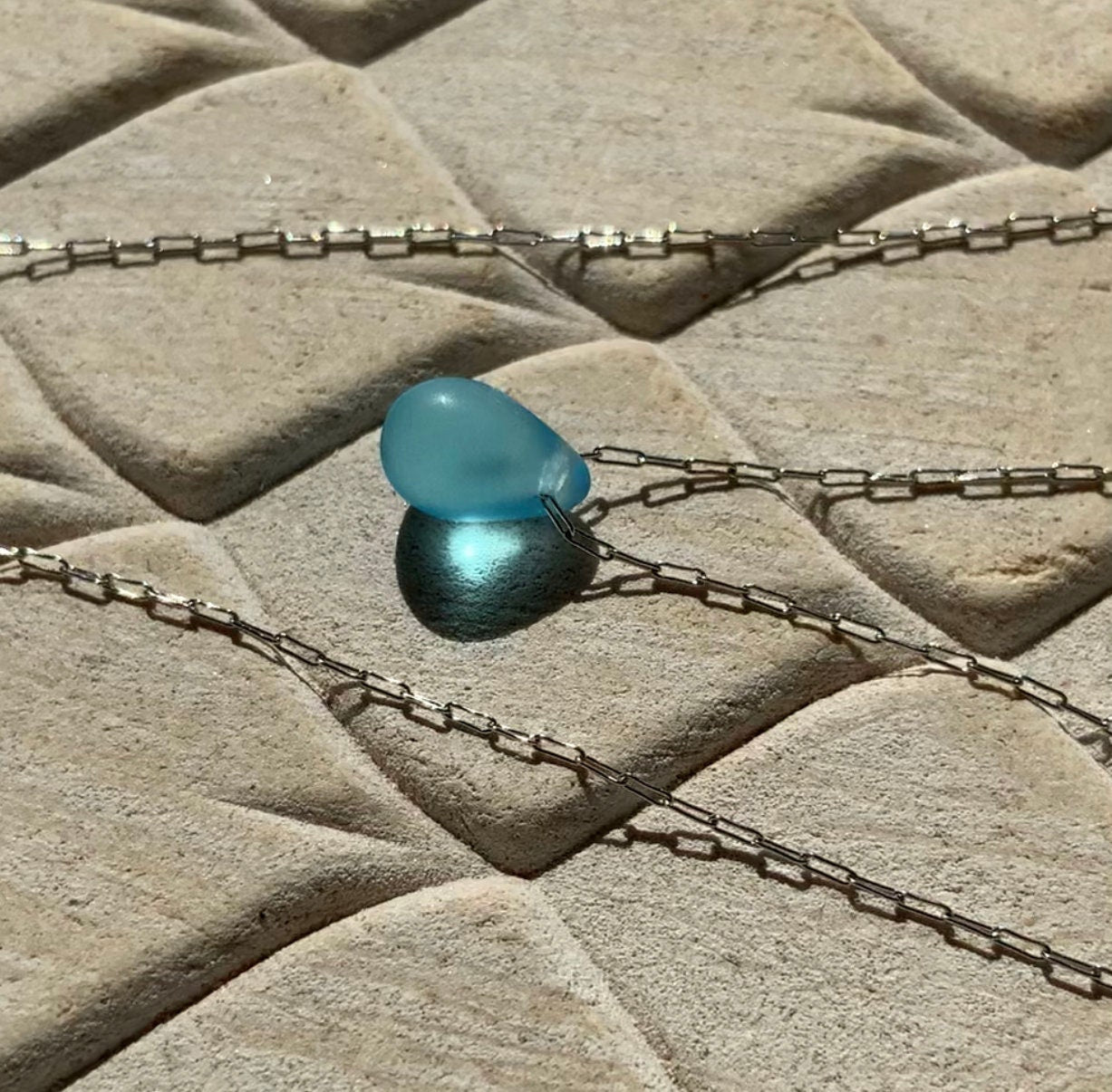 A vibrant aqua matte teardrop-shaped glass pendant, elegantly suspended on a stainless steel chain, styled on a beige textured background with natural sunlight enhancing its color
