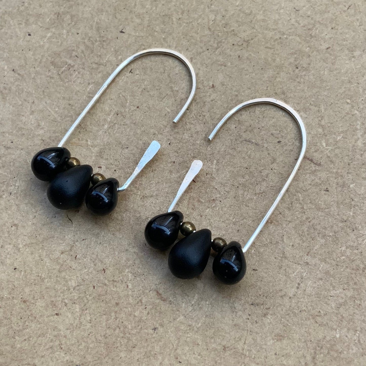 Close of minimalist silver hoop earrings with three smooth black teardrop glass pendants and brass bead accents, displayed on a wooden background for a contemporary and refined presentation.