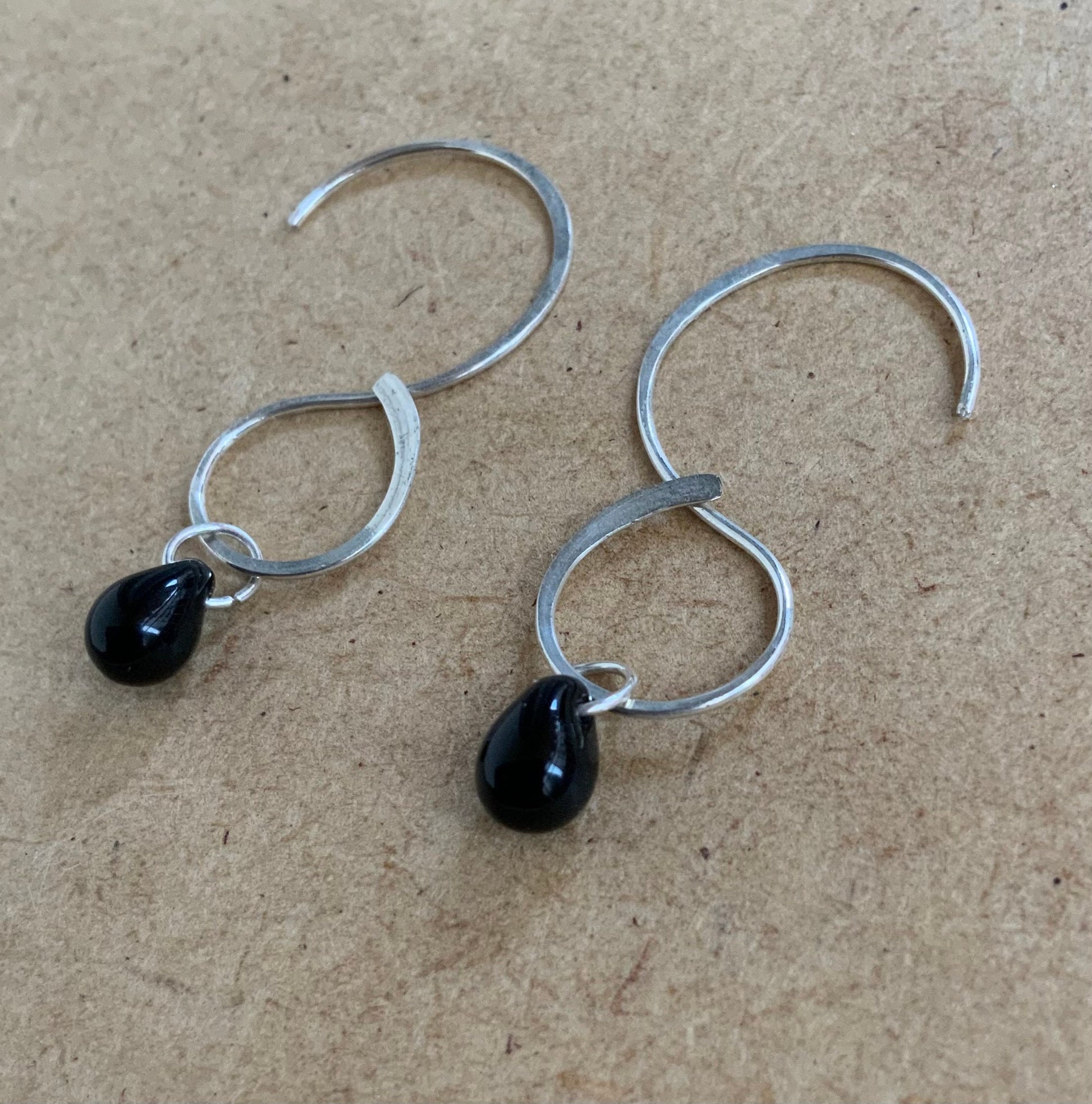 A pair of handcrafted infinity-style silver hoop earrings featuring black teardrop glass pendants, resting on a wooden surface. A sleek and modern design perfect for everyday elegance.