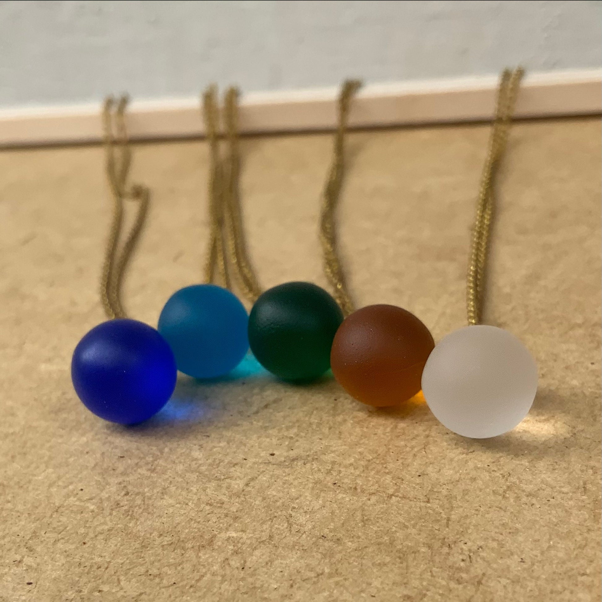 A close-up of matte teardrop glass pendant necklaces in vibrant shades of cobalt blue, turquoise, emerald green, amber, and frosted white, elegantly arranged on a wooden display. Handcrafted in Greece and timeless.