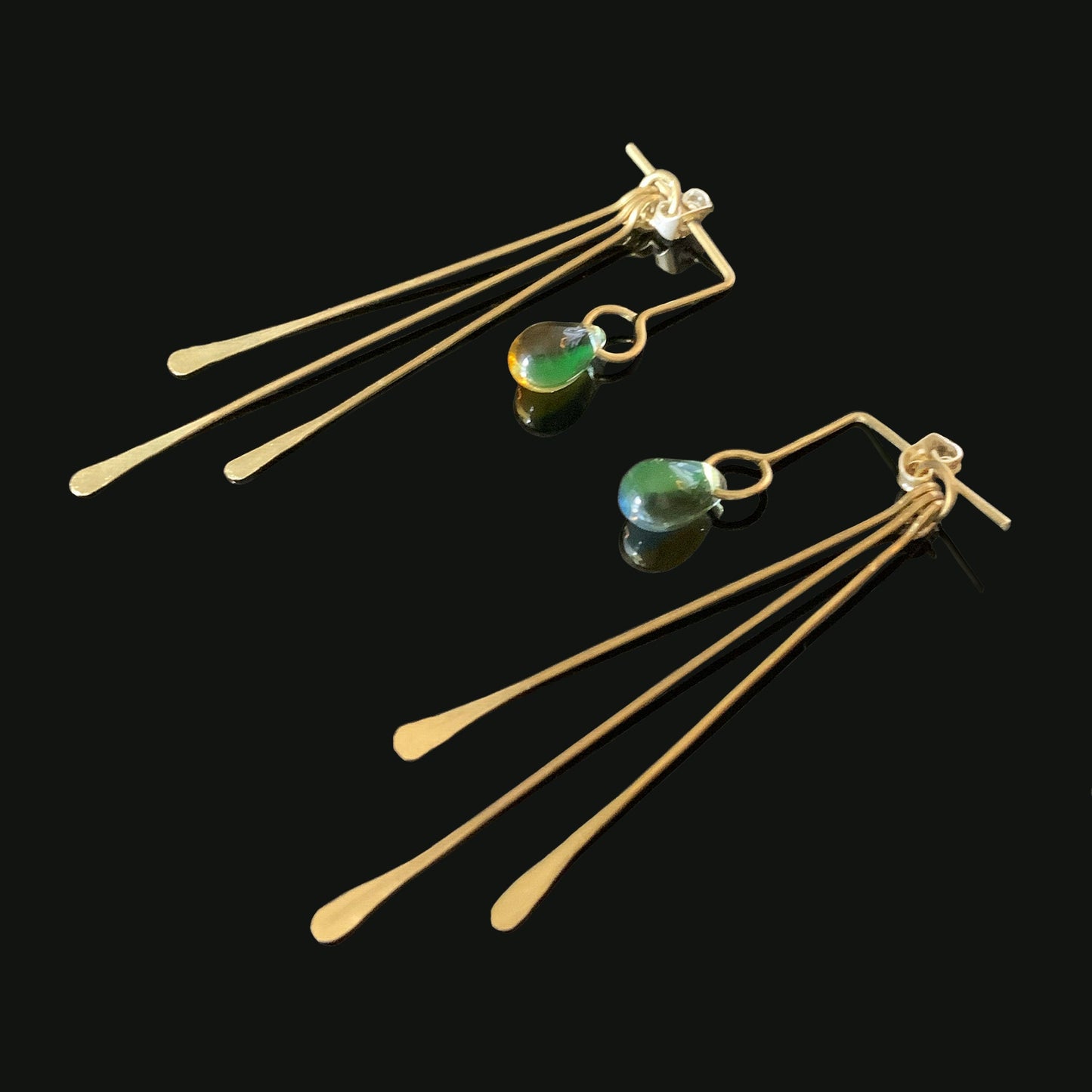 A pair of handcrafted brass earrings featuring gradient glass teardrop pendants in shades of green and amber, accented with elongated brass bars, displayed on a black background for a bold and modern look.