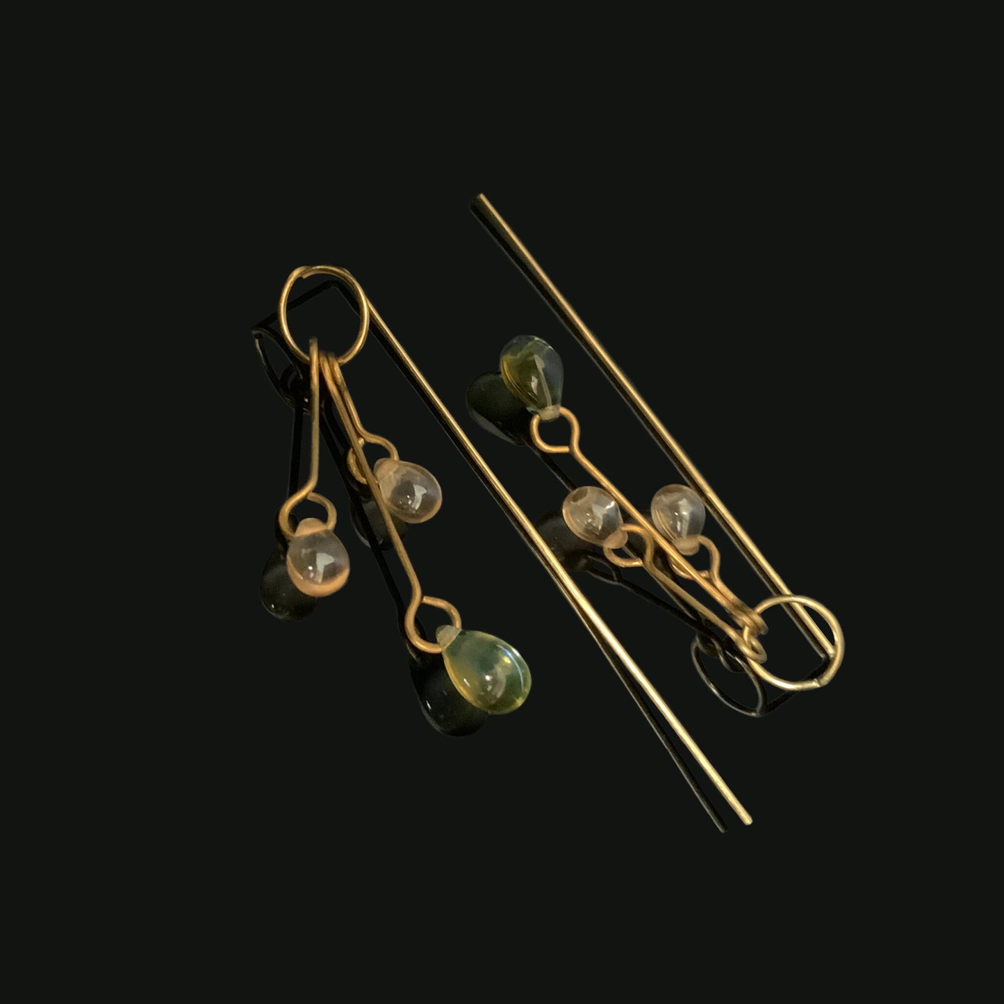 A pair of brass long dangle earrings adorned with clear and gradient glass pendants in soft hues of green and blue, styled on a black reflective background to emphasize their elegant design.