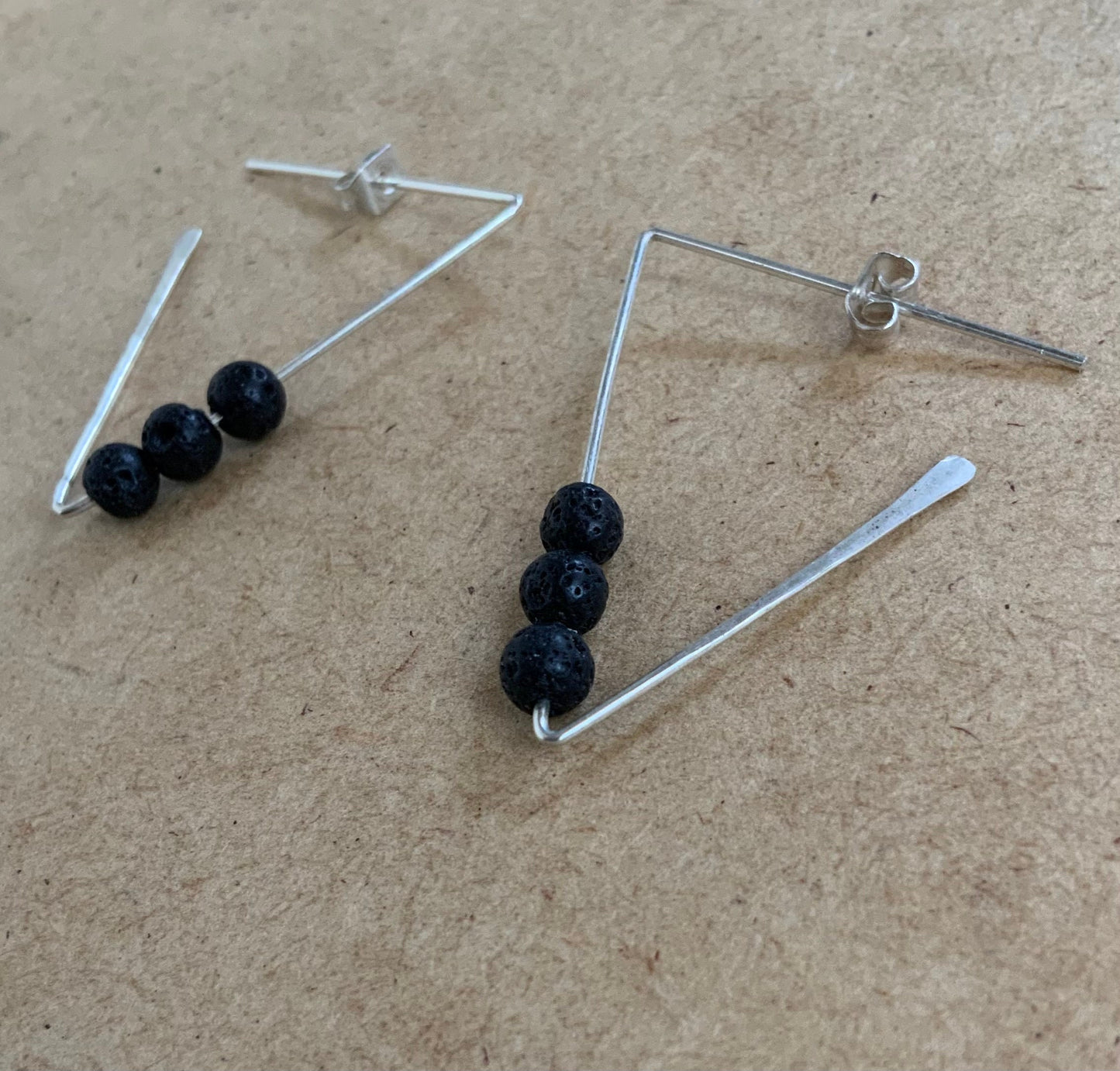 A close-up of silver geometric earrings featuring three textured black lava beads, resting on a wooden surface. A contemporary accessory with natural volcanic elements and hammered edges.