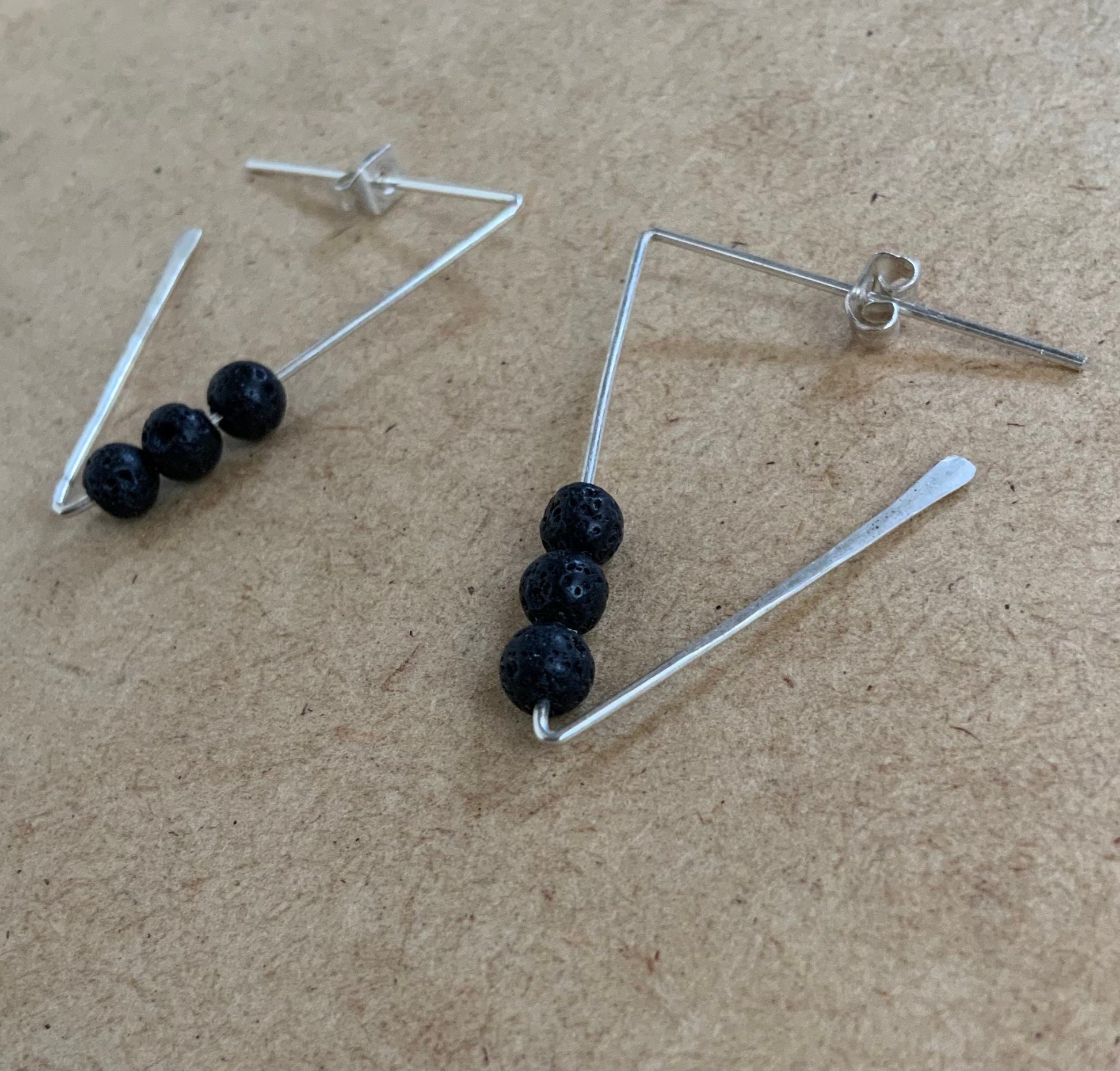 A close-up of silver geometric earrings featuring three textured black lava beads, resting on a wooden surface. A contemporary accessory with natural volcanic elements and hammered edges.